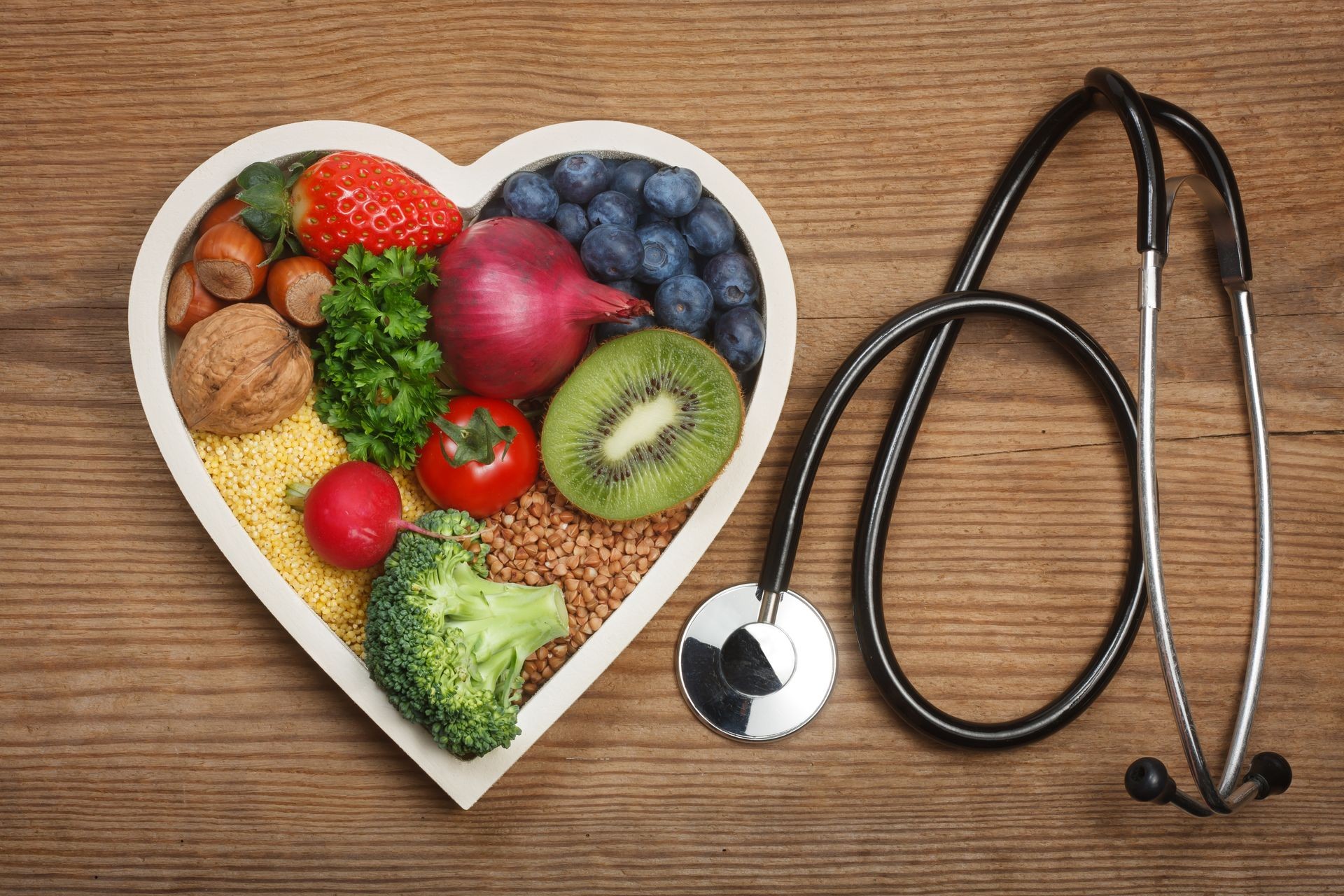 Healthy food in heart shaped bowl and stethoscope. Food such as blueberries, red onion, strawberry, parsley leaves, hazelnuts, walnut, tomato. kiwi, millet, buckwheat, radish, broccoli.